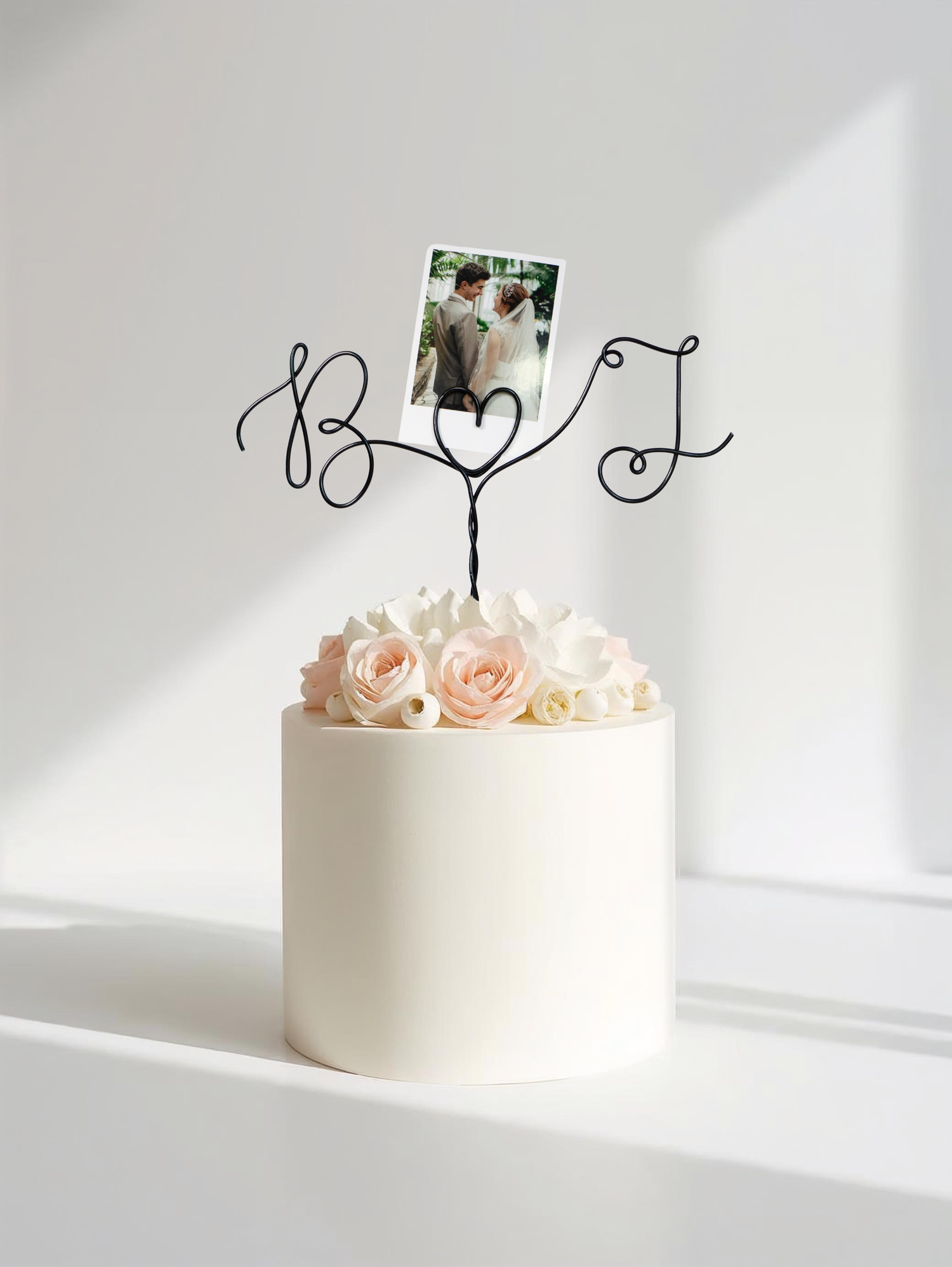 White cake with pink roses and a decorative monogram on top against a white background with wire cake topper with custom initials
