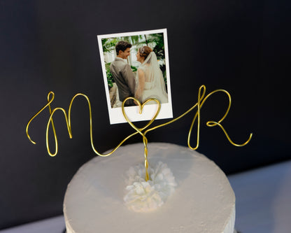 Gold wire cake topper with personalized initials on a cake against a dark background